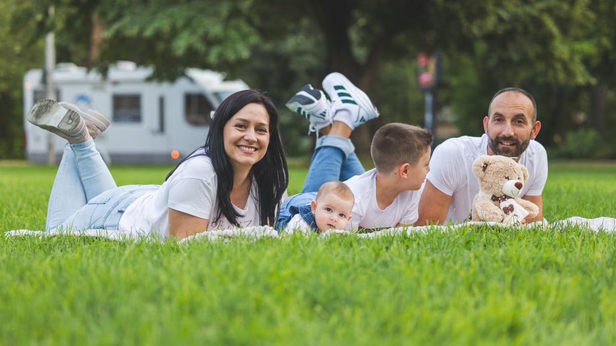 Happy family lying on grass with RV in the background, enjoying a sunny outdoor picnic.